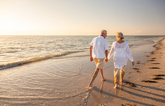 two elderly people walking on the beach next to the waves holding hands.