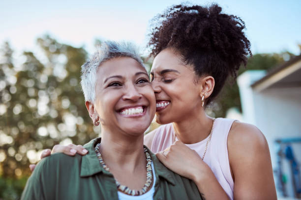 african american mom in a green shirt and daughter in a white shirt smiling at each other with trees and the sky in the background